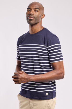 Man staat met gevouwen handen, draagt een marineblauw-wit gestreept T-shirt en beige broek tegen een effen achtergrond.