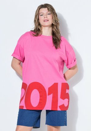 Kobieta w oversize różowym t-shircie z dużym czerwonym nadrukiem "2015" i podwiniętymi rękawami, zestawiona z niebieskimi, dżinsowymi szortami do kolan.