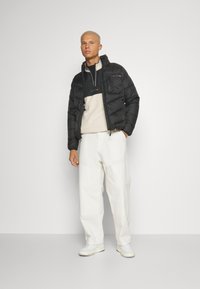 Svart dunjakke med hette og frontglidelås, beige fleece-lag under, løse hvite bukser og hvite sneakers.