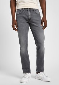 Hombre vestido con jeans gris ajustados y zapatillas blancas, de pie contra un fondo claro y liso.