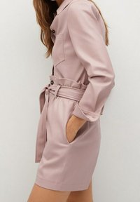 Jasnoróżowa sukienka z paskiem, z długimi rękawami, zapinana na guziki z przodu, z bocznymi kieszeniami i dopasowaną talią; gładki materiał o minimalnej teksturze.