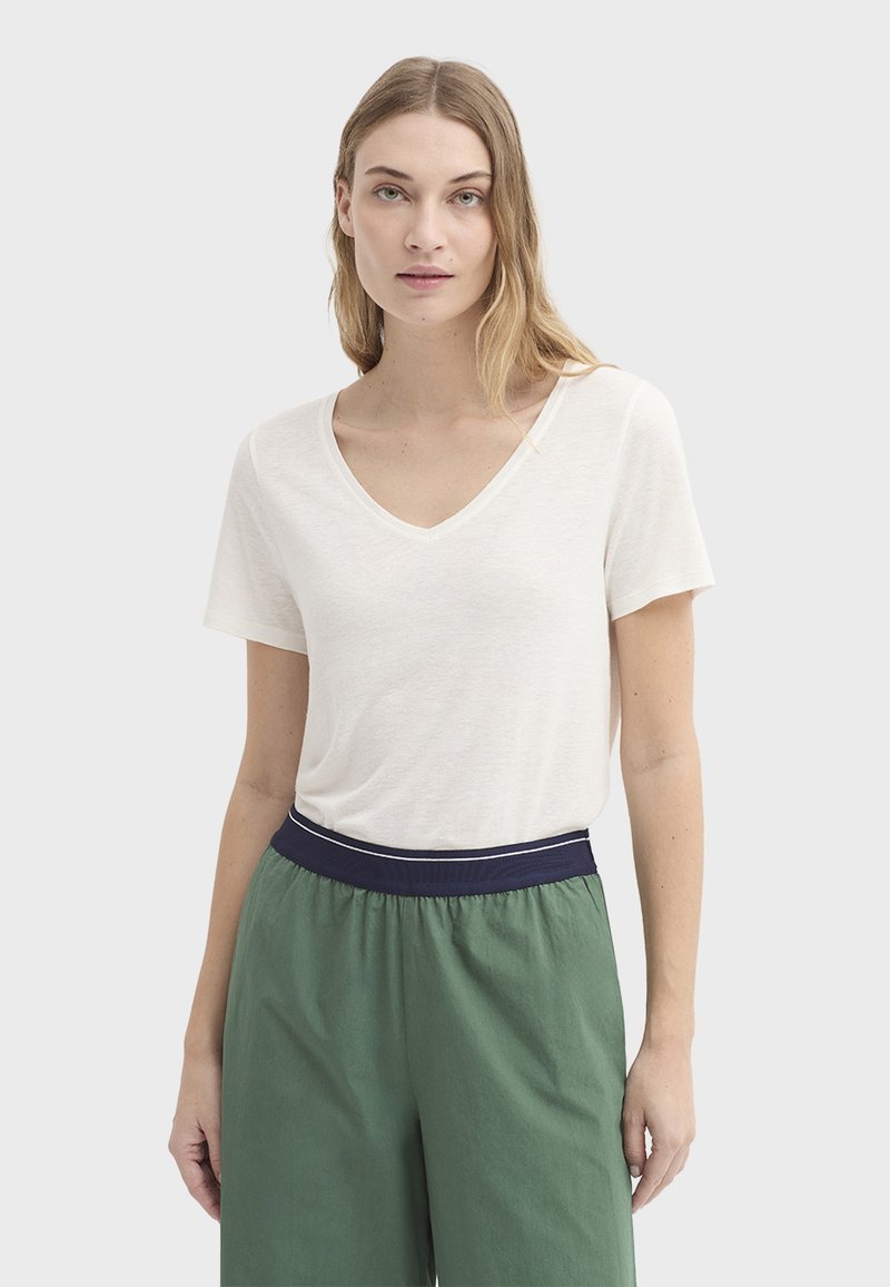 T-shirt à col en V et manches courtes en tissu beige clair. Associé à un pantalon vert avec une ceinture élastique bleu marine. Texture lisse.