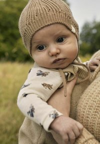 En baby i en stickad beige mössa och krämfärgad outfit med djurmönster, hålls nära av en vuxen klädd i en beige stickad tröja utomhus.