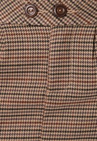 Brązowa i beżowa tkanina w kratę z wzorem pepitki, z dwoma ciemnymi guzikami na pasku i widocznymi kieszeniami.