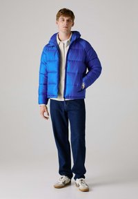 Blå pufferjakke med hætte, med quiltede sektioner og lynlåslukning, parret med mørk denimjeans og hvide sneakers.