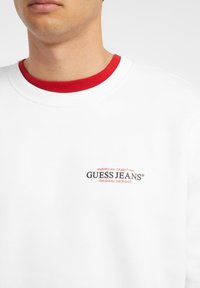 Hvit genser med rød ribbestrikket krage, med et trykket logo i svart og rødt tekst på forsiden. Glatt tekstur, uformelt design.