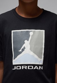 T-shirt en coton noir avec un grand motif blanc et gris représentant une silhouette en plein saut et le logo "JORDAN" en bas.