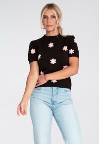 Kobieta ubrana w czarny sweter z krótkim rękawem w różowe wzory kwiatowe oraz jasnoniebieskie dżinsy, pozująca na tle jednolitego jasnego tła.