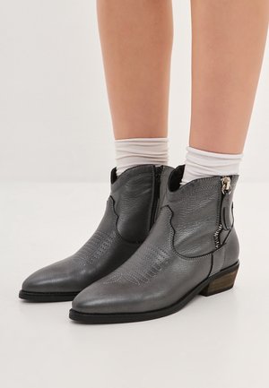 Bottines grises en cuir montant à la cheville avec fermetures éclair latérales portées avec des chaussettes blanches, montrées sur les mollets d'une personne contre un fond neutre.