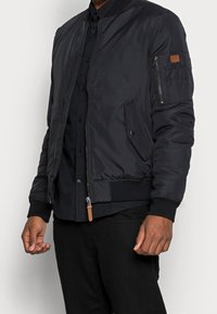 Bomber nero realizzato in tessuto liscio, con zip frontale, polsini a coste e dettagli con tasche su entrambi i lati e sulla manica sinistra.