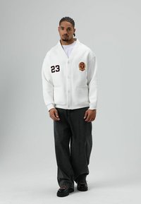 Witte gebreide cardigan met een sjaalkraag, knoopsluiting en bordeauxrode borduursels, gecombineerd met donkere brede jeans en zwart schoeisel.