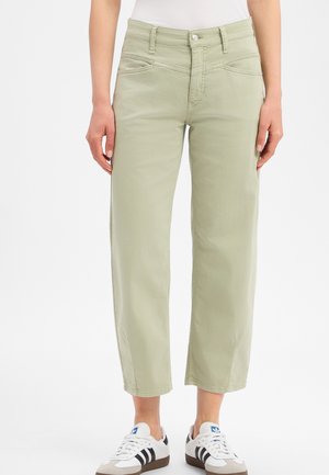 Persoon draagt lichtgroene cropped broek en witte sneakers met zwarte strepen, staand tegen een effen witte achtergrond.