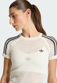 Camiseta blanca de punto con un patrón de malla, con rayas negras en los hombros y un logo bordado en el pecho. Mangas cortas.