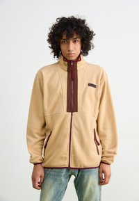 Beige fleece jacket with brown accents, high collar, front zipper, and two side pockets. The cuffs and hem have maroon trim for contrast.