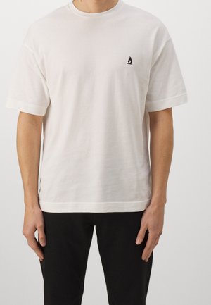 Personne portant un t-shirt blanc uni à manches courtes avec un petit logo de flamme noire sur la poitrine gauche, associé à un pantalon noir.