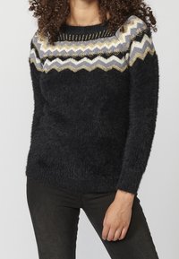 Frau trägt einen flauschigen schwarzen Pullover mit Zickzackmustern in Weiß, Gold und Grau auf der Brust und den Schultern, kombiniert mit schwarzen Hosen.
