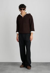 Donkerbruin gebreid vest met een witte kraag, gedragen met zwarte broek en bruine schoenen. Het vest heeft een rits en een ontspannen pasvorm.