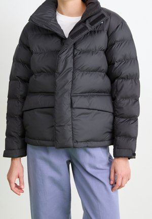 Person wearing a black puffer jacket with front pockets and a partially zipped collar over a white shirt and light blue pants.