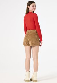 Maglia a maniche lunghe rossa a coste abbinata a pantaloni corti in velluto marrone a vita alta con tasche posteriori. Modella indossa stivaletti alla caviglia di colore avorio.