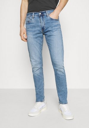 Homme portant un jean slim bleu clair avec une main dans la poche et des baskets blanches, debout devant un fond uni.