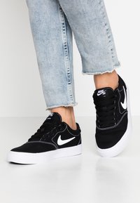 Baskets en toile noires avec accents blancs, présentant un design à lacets, un logo Nike blanc et une semelle en caoutchouc blanche. Tige texturée avec des détails de couture.