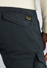 Dunkelblaue Cargo-Hosen mit einer Gesäßtasche mit einem gelben und schwarzen Markenlabel, strukturiertem Stoff und einer sichtbaren Seitentasche.