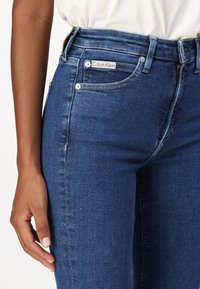 Pravá ruka odpočívající po boku, oblečené tmavě modré džíny Calvin Klein a bílou košili, viditelné švy džínů a detail přední kapsy.