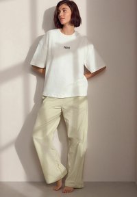 Vit överstor t-shirt med texten "PARIS", i kombination med löst sittande byxor i ljusgul rutig design. Modellen står barfota.