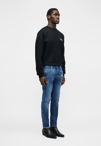 Άντρας μοντέλο που φοράει μαύρο μακρυμάνικο φούτερ, εφαρμοστά μπλε τζιν και μαύρα δερμάτινα μποτάκια μέχρι τον αστράγαλο, στέκεται μπροστά σε απλό λευκό φόντο.