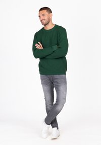 Grüner Strickpullover mit strukturiertem Muster, kombiniert mit hellgrauen, verwaschenen Jeans und weißen Turnschuhen. Das Model steht mit verschränkten Armen.