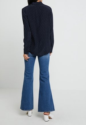 Femme portant une chemise à manches longues bleu marine à pois, un jean évasé bleu et des sandales blanches à talons, debout devant un fond uni.