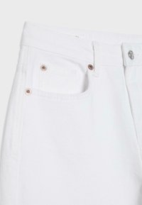 Gros plan sur un jean en denim blanc montrant une poche avant avec des rivets, des passants de ceinture et une fermeture à bouton métallique à la taille.