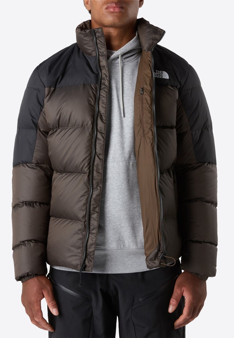 Mann in einer schwarz-braunen Puffjacke über einem grauen Hoodie und schwarzen Hosen, stehend vor einem schlichten Hintergrund.