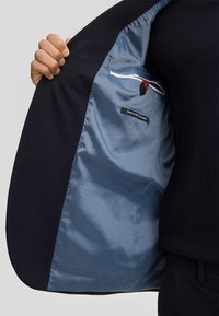 Blazer nero, interno foderato con tessuto azzurro chiaro. Presenta un'etichetta, un bottone e cuciture a contrasto. Texture liscia, design aderente.