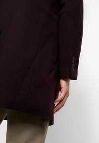 Vue de côté d'une personne portant un manteau violet foncé avec quatre boutons noirs sur la manche et un pantalon vert olive sur fond blanc.