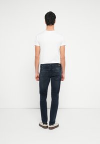 Młody mężczyzna stojący tyłem, ubrany w białą koszulkę, ciemnoniebieskie dżinsy i białe trampki na jednolitym białym tle.