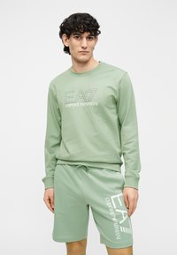 Sudadera de algodón verde claro con mangas largas y cuello redondo; presenta el logo EA7 en bordado contrastante. Pantalones cortos a juego con el logo.