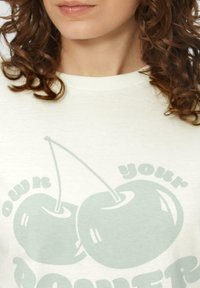 Lichtgekleurde katoenen T-shirt met een afbeelding van twee groene kersen en de tekst "own your power" in stijlvol lettertype.