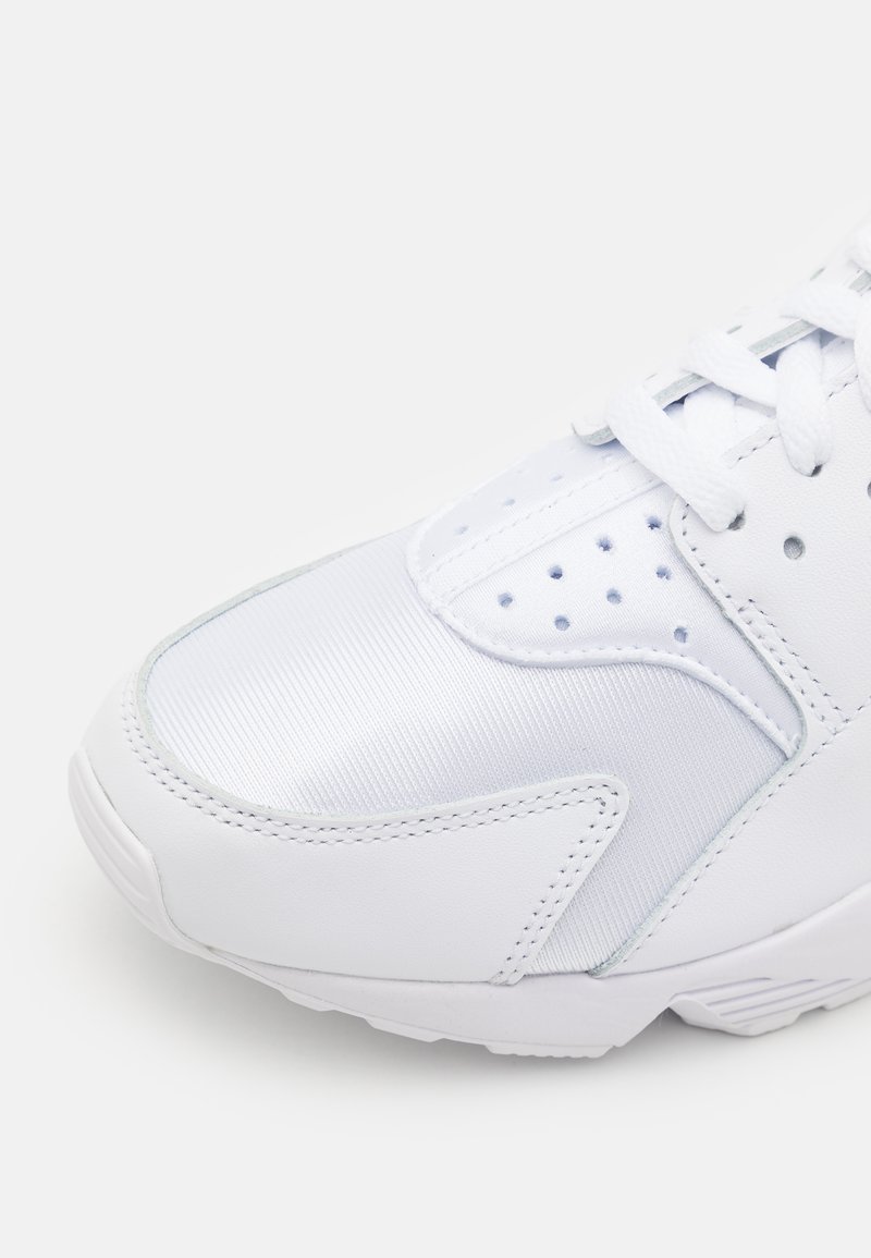 Weißer Sport-Schuh mit einer Kombination aus glattem Leder und strukturierter Textiloberfläche. Er verfügt über perforierte Details, flache Schnürsenkel und eine gepolsterte Sohle.