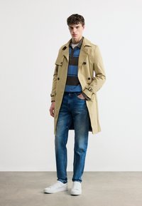 Νεαρός άνδρας φοράει μπεζ καμπαρντίνα πάνω από μπλε και καφέ ριγέ πόλο, μπλε τζιν και λευκά αθλητικά παπούτσια, στέκεται με το ένα χέρι στην τσέπη.