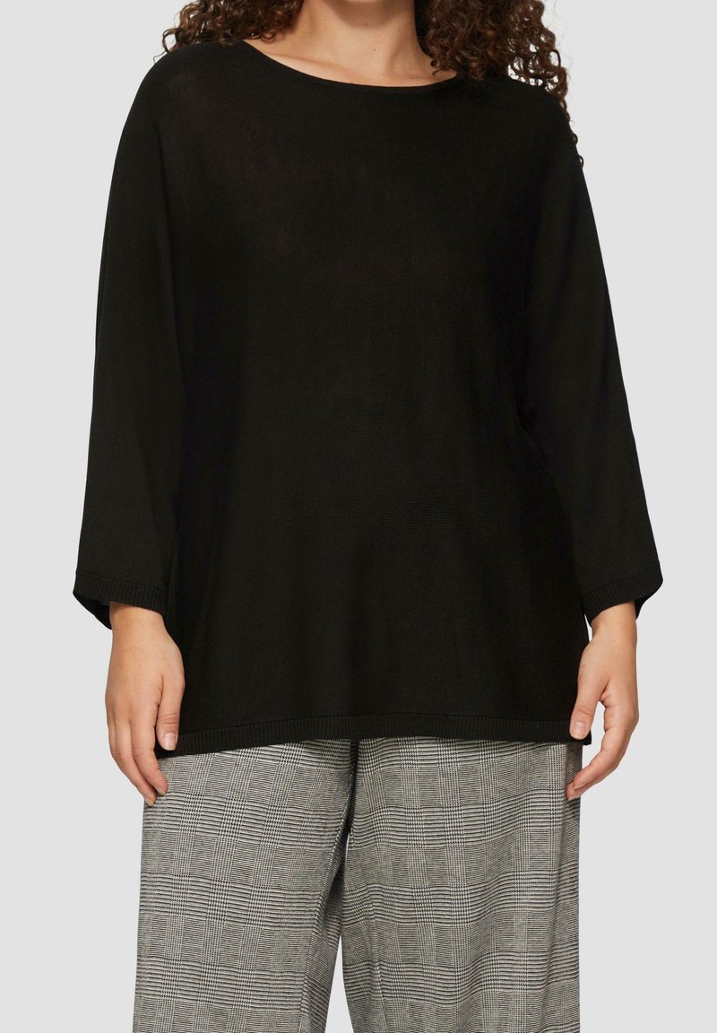 Schwarzer Strickpullover mit weitem Ausschnitt und Dreiviertelärmeln, kombiniert mit grauen Karo-Breitehosen. Strukturiertes Gewebe sichtbar.