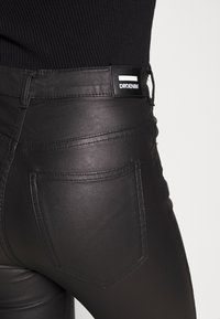Calças skinny em imitação de pele pretas com uma cintura alta, apresentando um design de bolso traseiro e um rótulo de marca na cintura. Textura suave.
