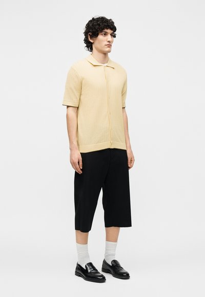 Νέος άνδρας με σγουρά μαλλιά που φοράει μπεζ κοντομάνικο πουκάμισο με κουμπιά, μαύρο φαρδύ κοντό παντελόνι, λευκές κάλτσες και μαύρα μοκασίνια, όρθιος.