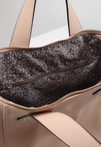 Jasnoróżowa skórzana torba typu tote z brązowym wnętrzem z materiału w kwiatowy wzór, wyposażona w kieszeń na zamek i metalowe ozdobne elementy.