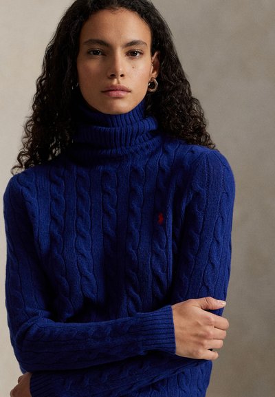 Blå kabelstrikket genser med høy hals og ribbestrikkede mansjetter og krage. Har en liten rød logo på brystet. Stoffet ser mykt og teksturert ut.