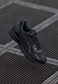 Chaussure de sport noire avec un dessus texturé, dotée d'un design à lacets, d'un logo distinctif et d'accents réfléchissants sur les côtés.