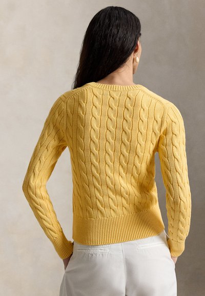 Femeie cu păr lung și închis la culoare, purtând un pulover galben tricotat cu model și pantaloni albi, văzută din spate, pe un fundal neutru.