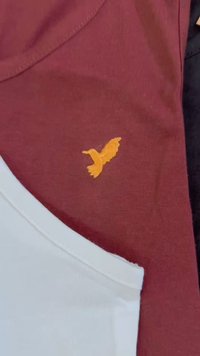 Camiseta de tecido bordeaux com um decote arredondado, apresentando um logótipo de pássaro bordado laranja no peito; camisa branca parcialmente visível abaixo.