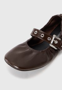 Ballerine plate en cuir marron avec des bords élastiques et une bride brillante ornée d'œillets métalliques et d'une boucle argentée sur fond blanc.