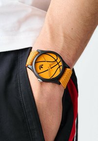 Reloj negro redondo con una esfera naranja brillante que presenta un diseño de baloncesto, complementado por una correa amarilla texturizada. Detalle de corona plateada visible.